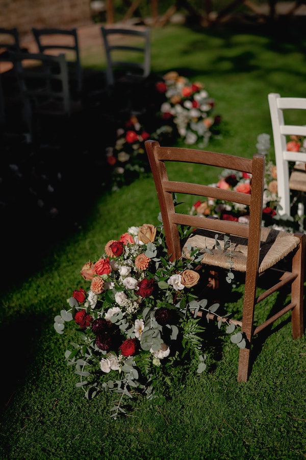 Colourful floral arrangement beside a wooden chair at an outdoor wedding ceremony